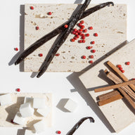 Vanilla beans, red peppercorns, and cinnamon sticks on a light stone surface.